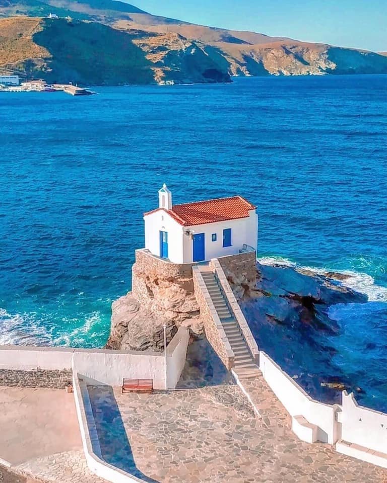 Scenic view of a traditional white chapel on a rocky islet in Andros, Greece, surrounded by deep blue Aegean waters.