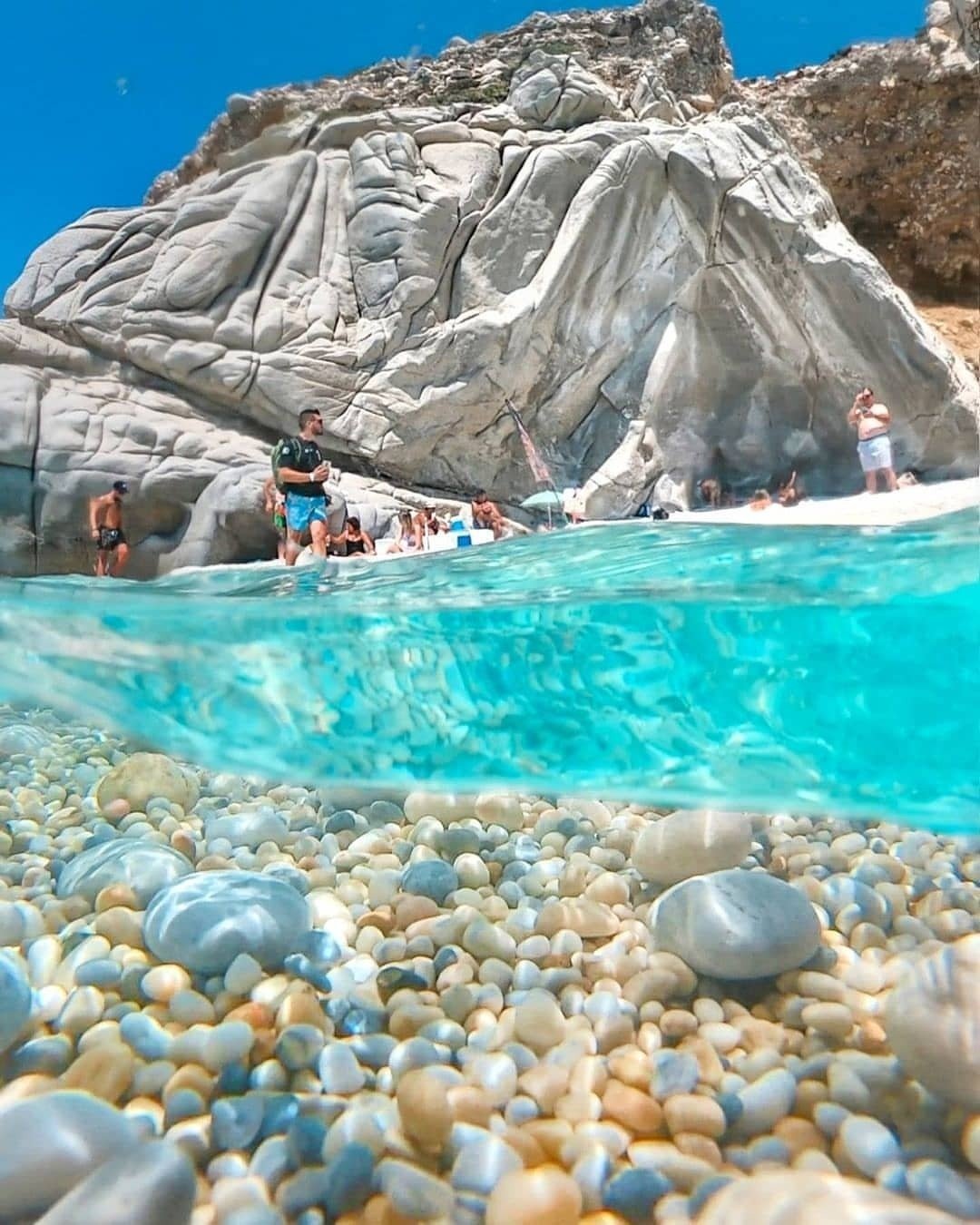 Crystal-clear turquoise waters with a split view of an underwater pebble beach and impressive rock formations on the coast of Ikaria, Greece.