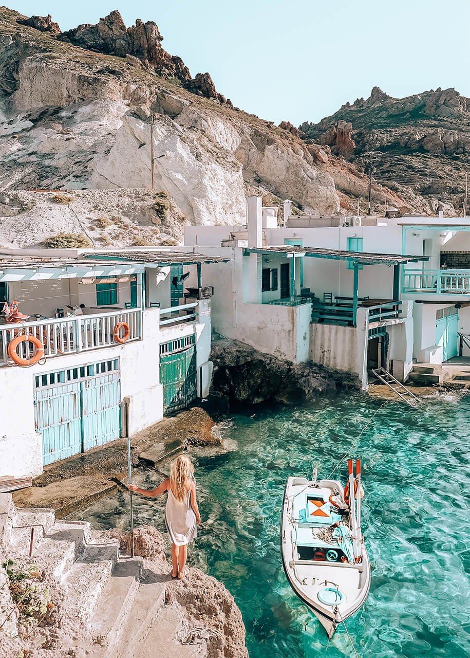 scenic fishing village in Milos, Greece, with traditional white and turquoise houses, a boat floating on crystal-clear waters, and a woman in a white dress enjoying the view.