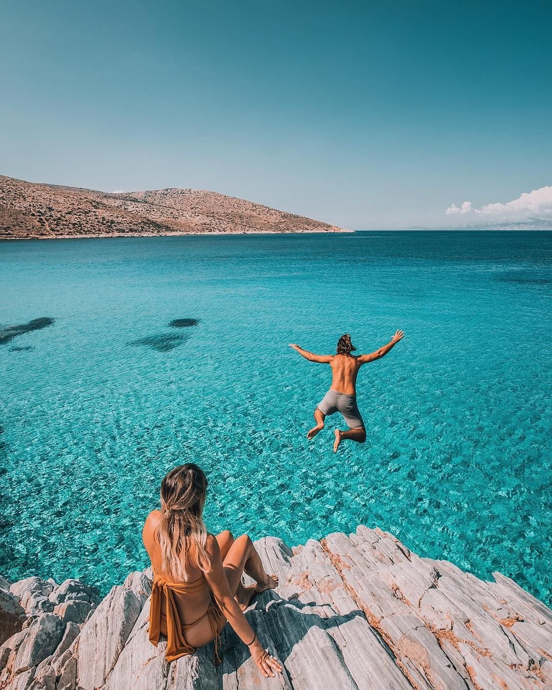 Man jumping into crystal-clear waters in Ios, Greece, while a woman in a bikini enjoys the view from rocky cliffs