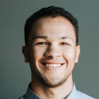 A smiling young man with short, curly dark hair, wearing a light blue shirt, set against a plain gray background.