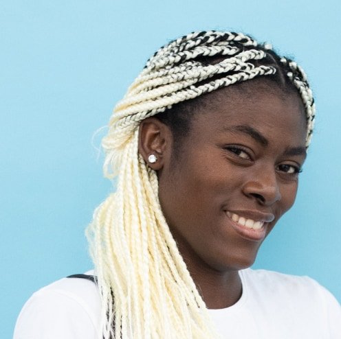 A smiling young woman with blonde braided hair styled in cornrows, wearing a white shirt, against a bright blue background.