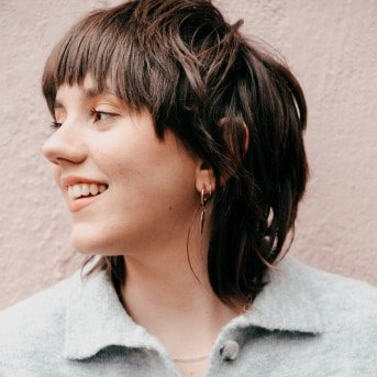 A young woman with short, layered dark hair and hoop earrings, smiling and looking to her left, set against a pastel pink background.